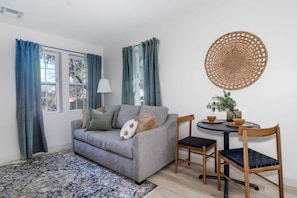 A living room with a grey sofa, two wooden chairs, a round table with a woven placemat, and a blue patterned rug.