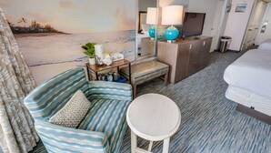 A hotel room with a beach mural, a sofa, a bedside table, a lamp, a TV, and a bed.