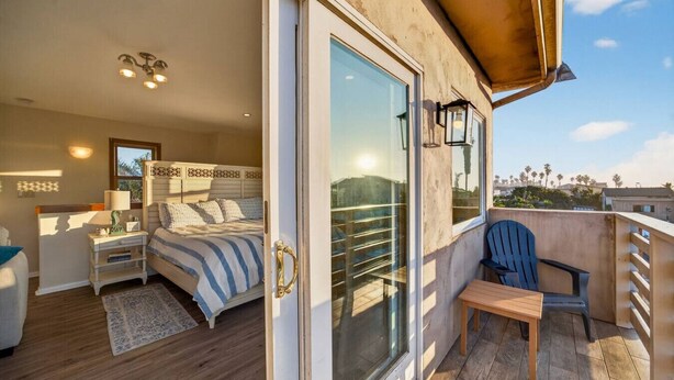 Une chambre avec un lit, une table de chevet, une lampe et une vue sur un balcon, accessible par une porte-fenêtre coulissante, où se trouvent une chaise et une table.