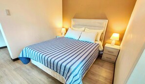 Una cama bien hecha con un edredón a rayas, dos lámparas de noche, una mesita de noche y un interruptor de pared.