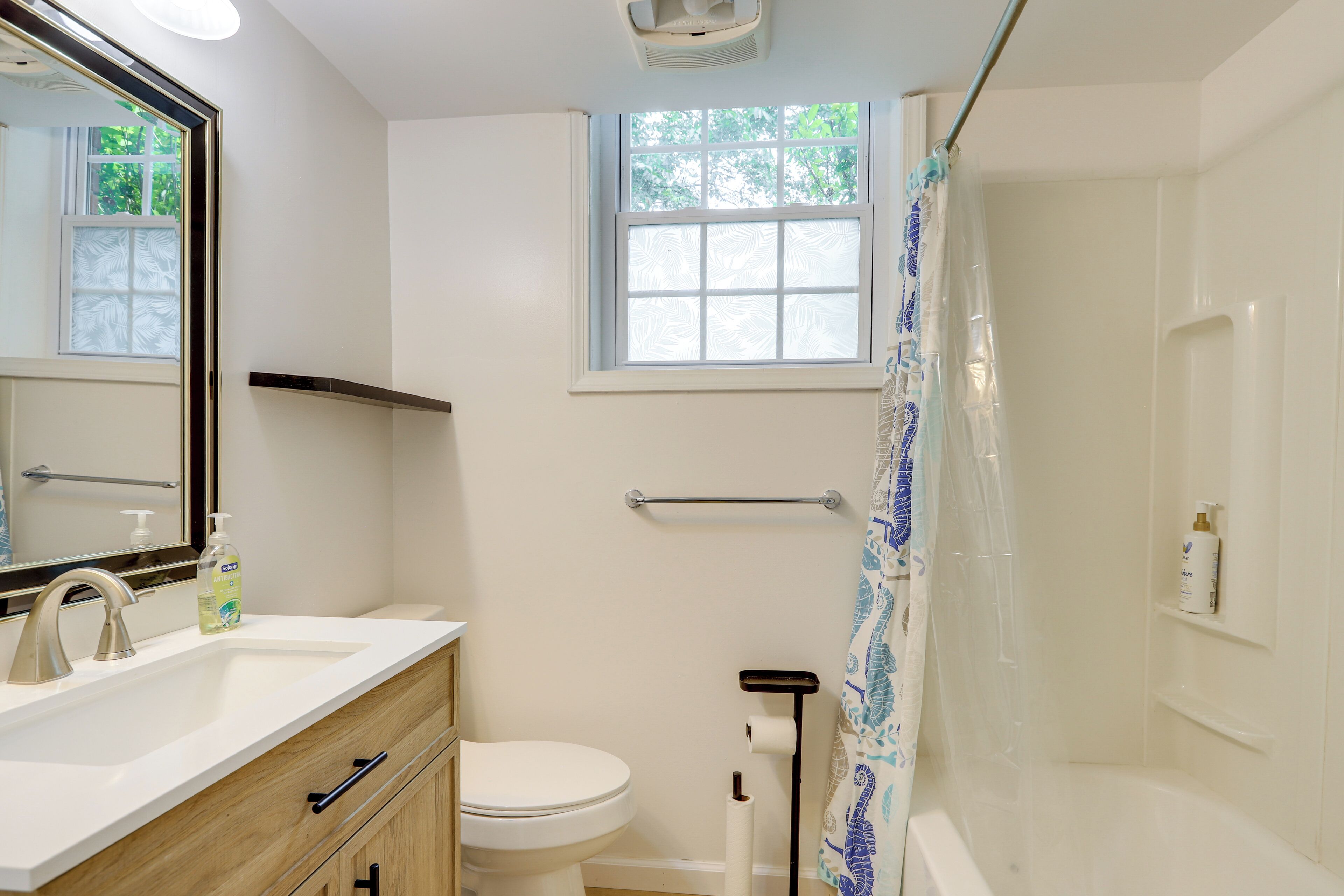 Un baño con lavamanos blanco, inodoro, ducha con cortina azul y blanca, ventana y espejo.