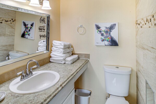 A bathroom with a sink, mirror, and toilet. There are towels stacked on the counter and two framed pictures of dogs on the wall. - Swim, Fish & Suntan: Cozy Corpus Christi Retreat (Corpus Christi)