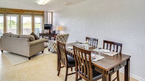 A dining area with a wooden table set for four, surrounded by wooden chairs. A sofa and a television are visible in the background. - Swim, Fish & Suntan: Cozy Corpus Christi Retreat (Corpus Christi)