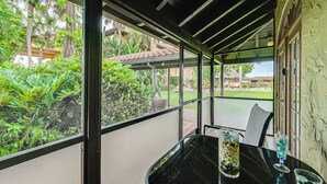A covered outdoor seating area with a table and chairs, surrounded by glass walls and a view of lush greenery.