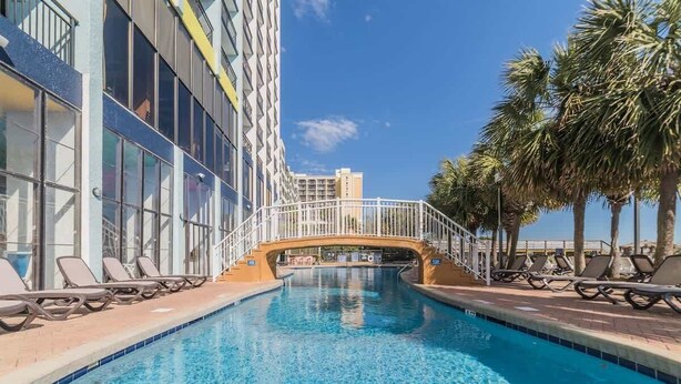 A swimming pool with lounge chairs and a bridge, surrounded by palm trees and modern buildings.