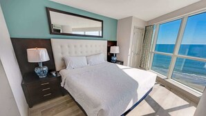 Un'ampia camera da letto con un letto grande, due comodini con lampade, uno specchio sopra il letto e una grande finestra con vista sull'oceano.