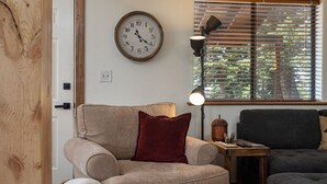 A cozy living room with a beige armchair, a dark grey sofa, a wooden floor, a wall clock, a table lamp, and a window with blinds. - Strawberry Summit Cabin Retreat (Duck Creek Village)