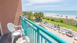 A balcony with a view of a beach, lounge chairs, and a pool area.