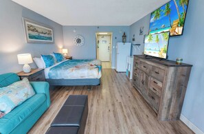 A bedroom with a bed, a nightstand, a dresser, a TV, and a door leading to another room. - New Direct Oceanfront at Sandcastle South | Pools! (Myrtle Beach)