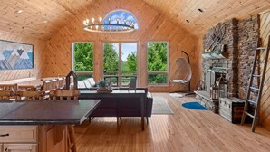 Interior acogedor de una cabaña con techo de madera, comedor, chimenea de piedra y ventanas amplias que dan a vistas de follaje.