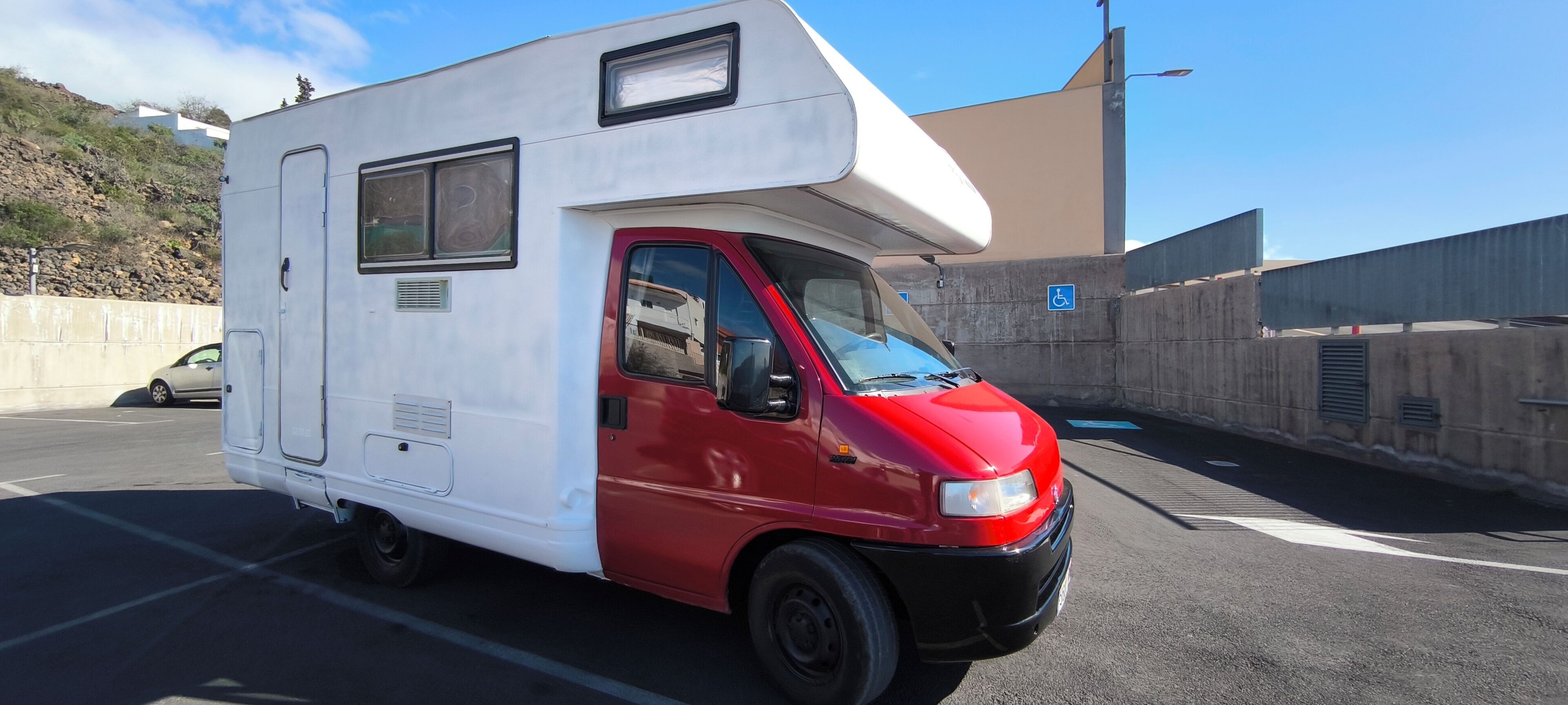 Un camping-car blanc et rouge est garé sur une route, avec une barrière en béton et, au fond, un panneau bleu indiquant un espace réservé aux personnes handicapées.