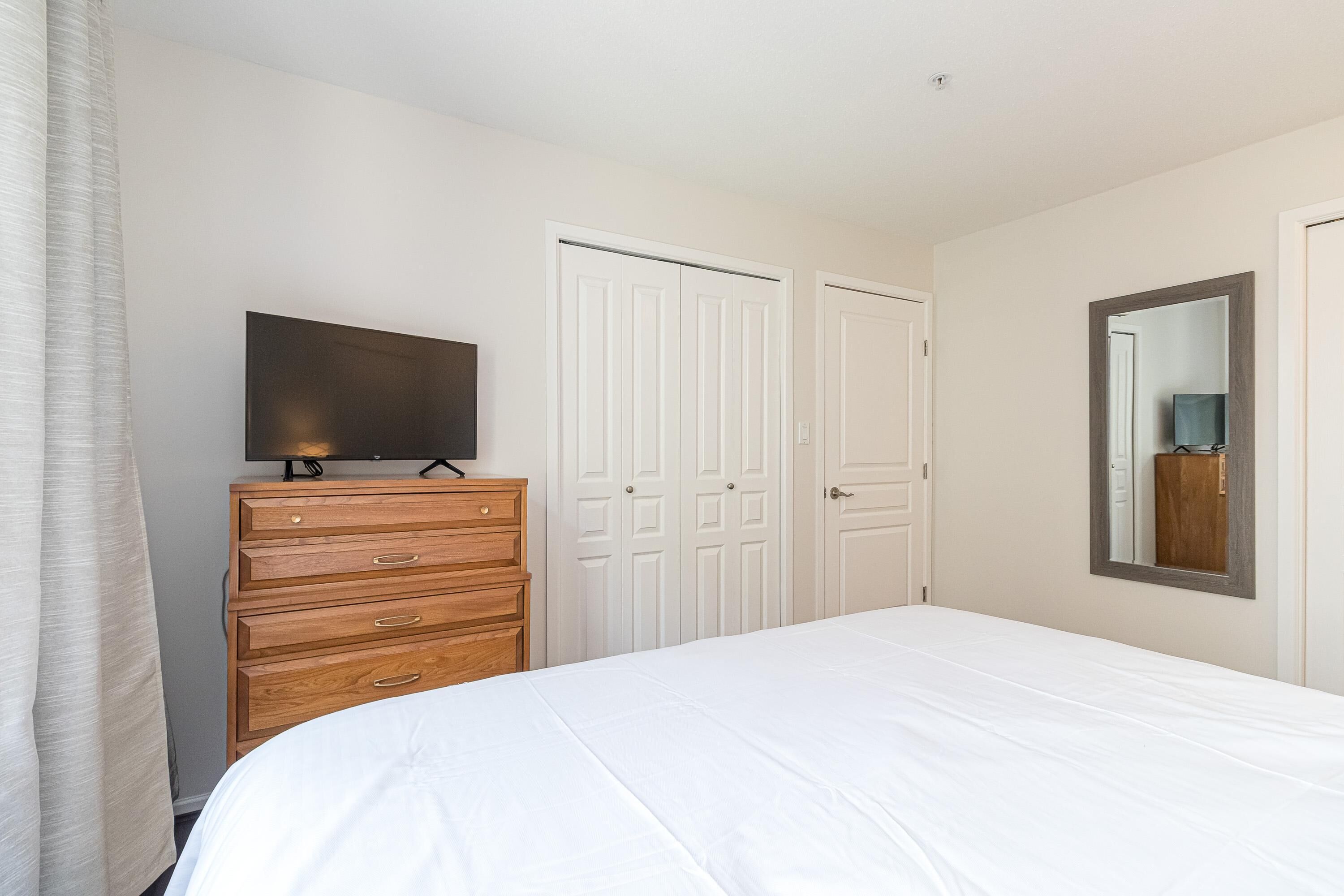 A bedroom with a bed, a wooden dresser, a flat-screen TV, white doors, and a mirror.