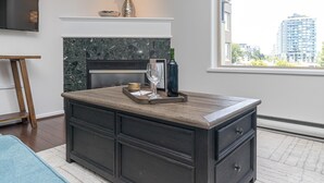 A modern living room with a dark wooden coffee table, a fireplace, and a large window offering a city view. - Waterfront Condo and Walk to Town/Trails (Esquimalt)