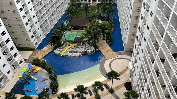 A high-rise hotel with a central swimming pool, a children's play area, and a landscaped courtyard with palm trees. -  Shore Residences MOA by darpm (Pasay)