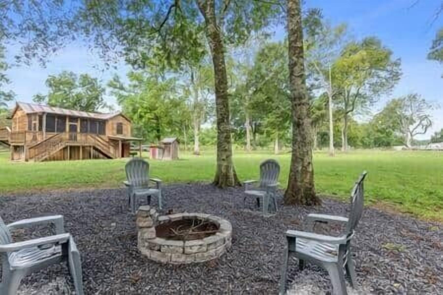 A rustic outdoor seating area with a fire pit, surrounded by trees and a wooden building in the background.