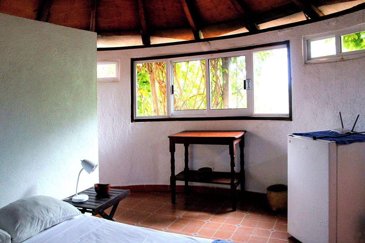 A bedroom with a bed, a desk, a small table, a window, and a refrigerator.