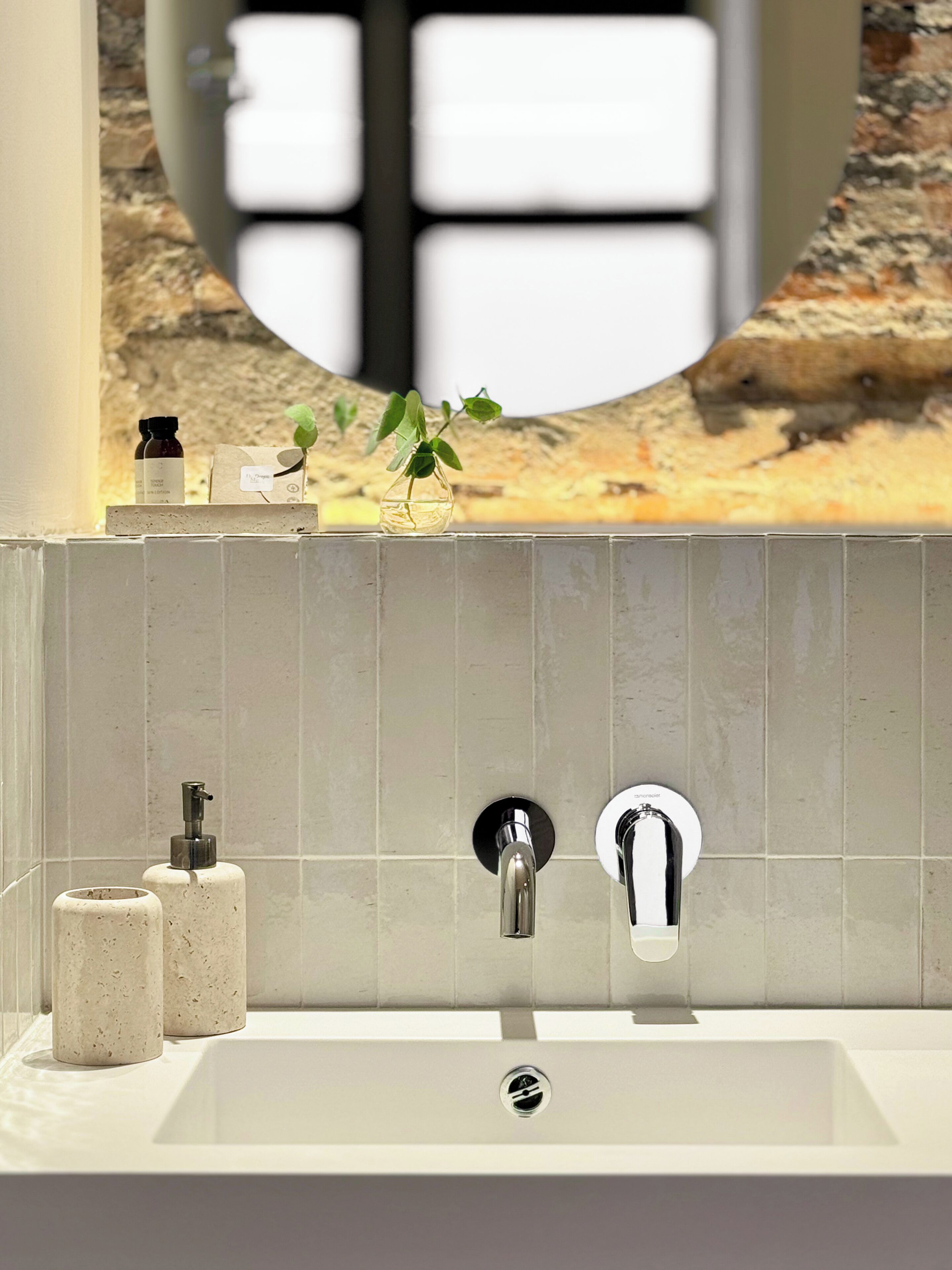 Et badeværelse med en hvid vask, to haner og et spejl. Der er toiletartikler på bordet og en lille plante på vasken.