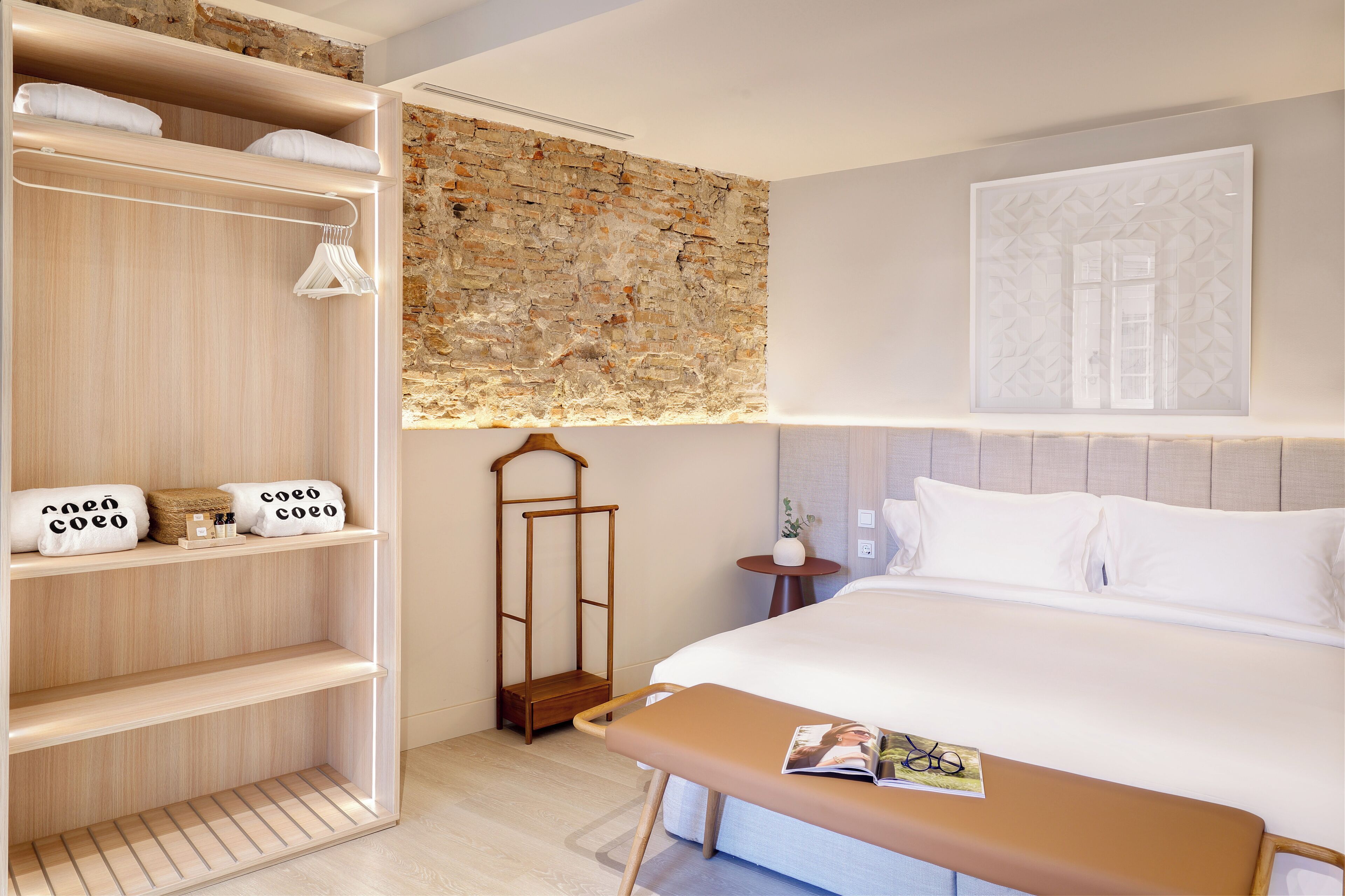 Quarto de hotel com uma cama grande, um guarda-roupa de madeira, uma mesinha com uma revista e uma decoração na parede.