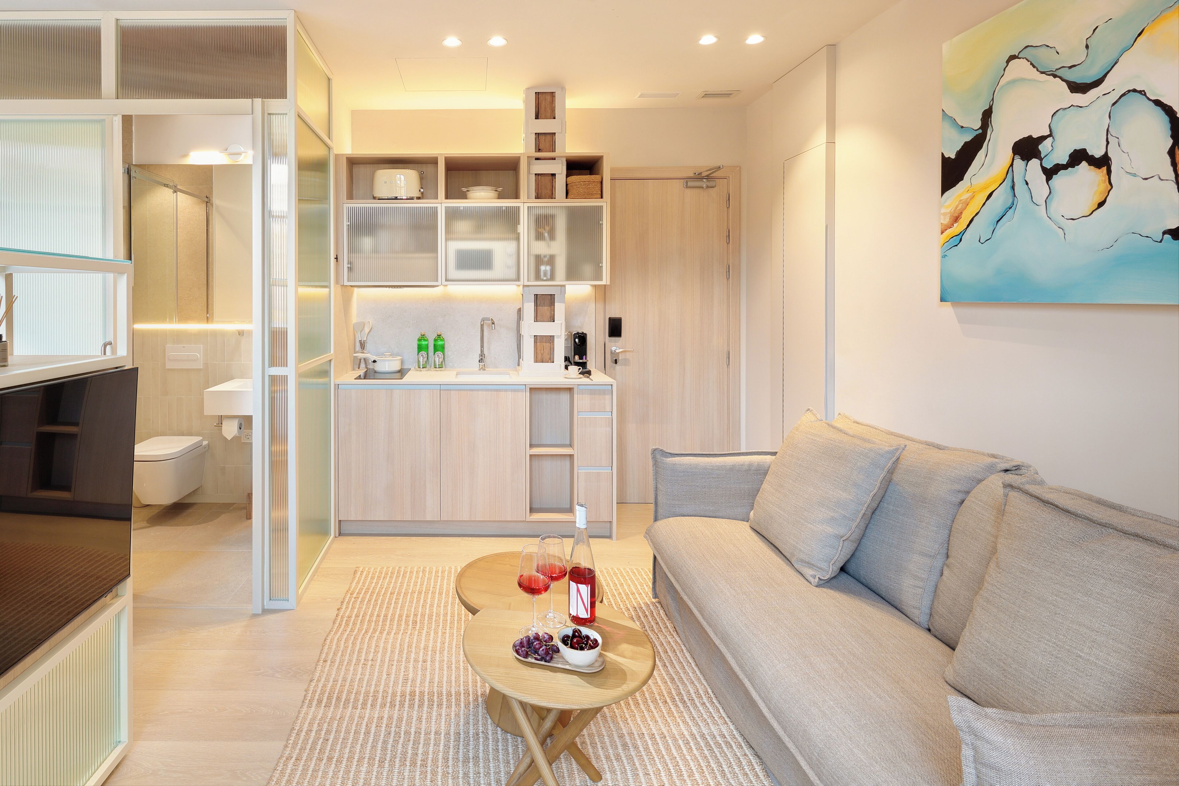 Una sala de estar moderna con un sofá, una mesa de centro con una botella y vasos, y un área de cocina con microondas y armarios.