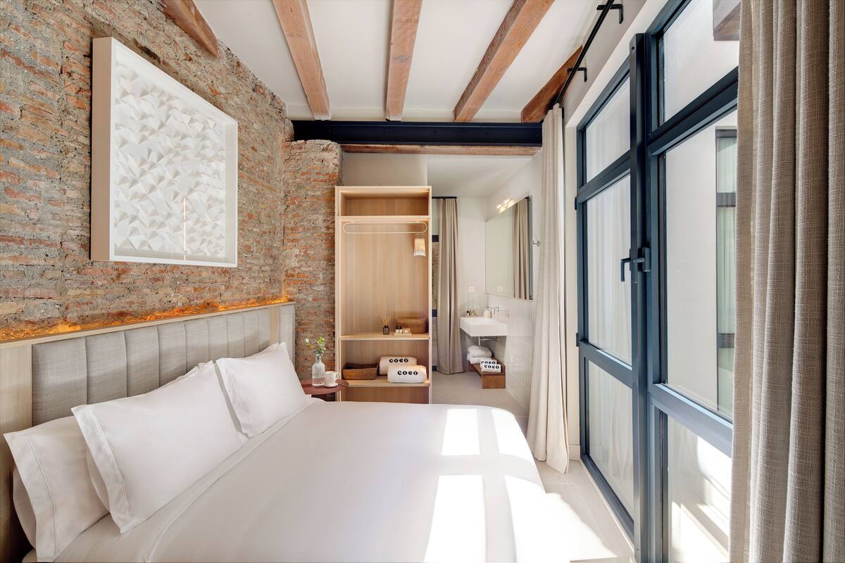 A modern bedroom with a brick wall, a large bed, a wooden wardrobe, and a bathroom visible through an open door.