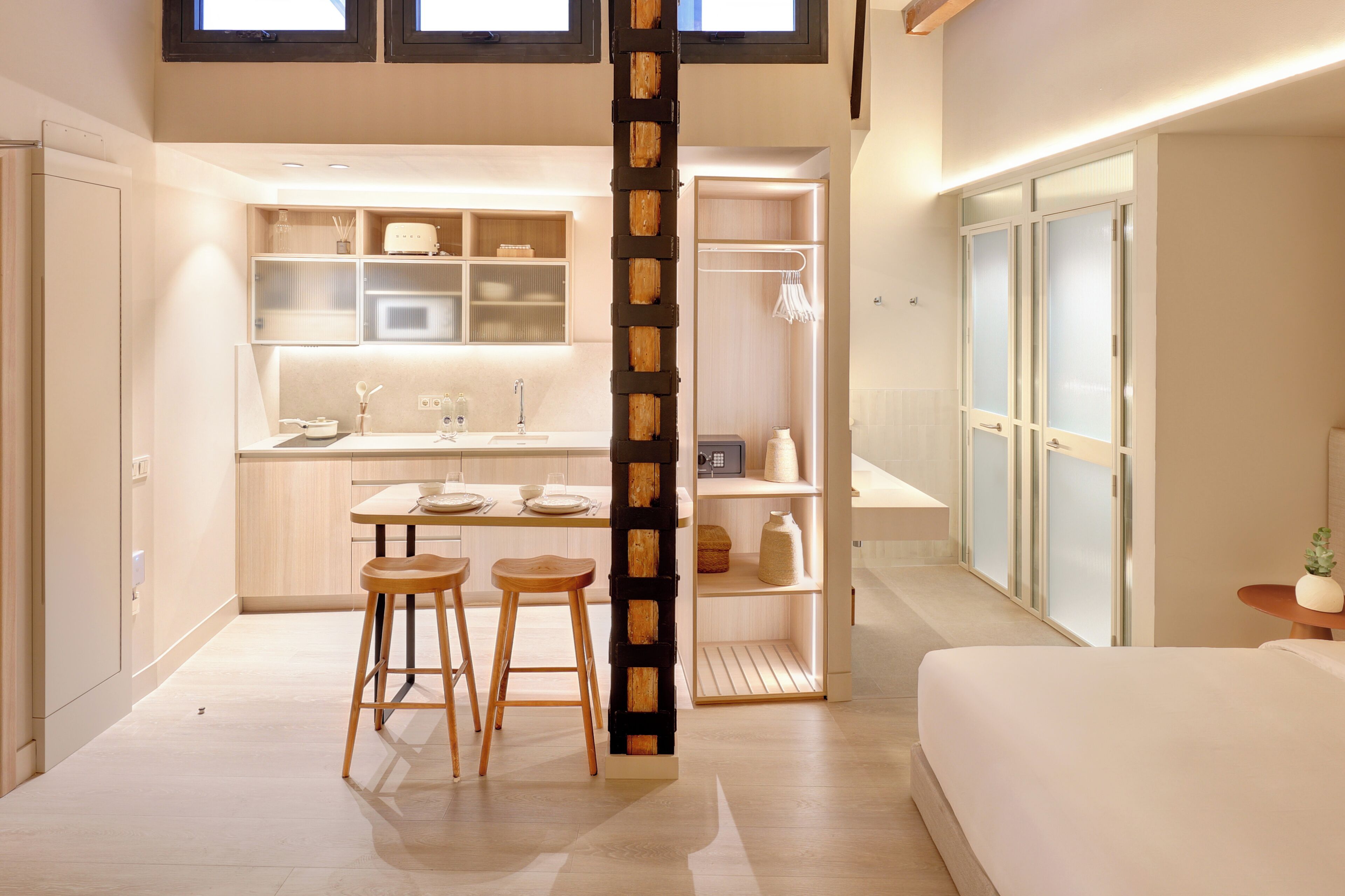Habitación de hotel compacta con kitchenette, zona de comedor con taburetes, cama y una pequeña mesa con una planta.