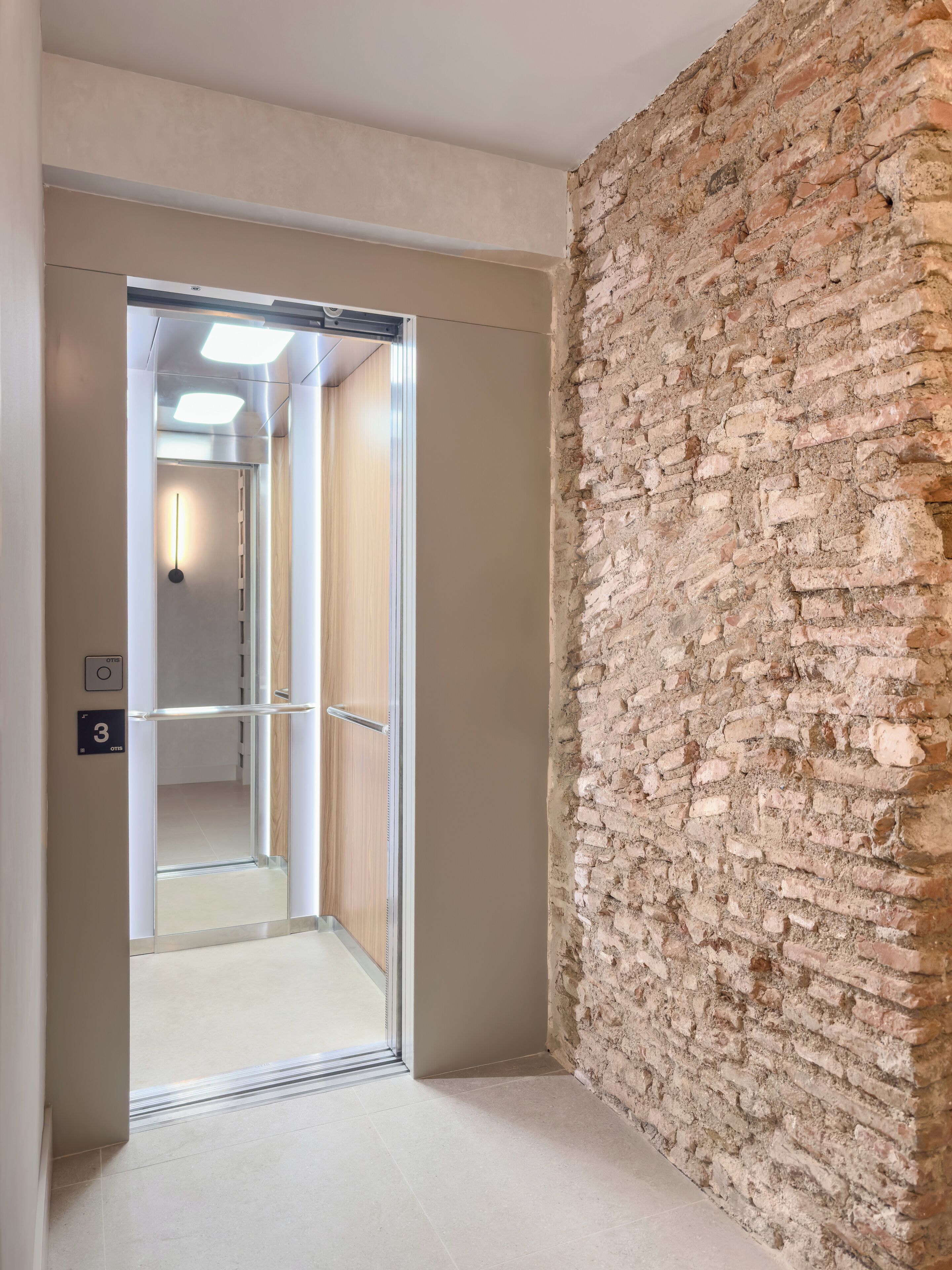 A hallway with a modern elevator, a brick wall, and a door leading to a room.