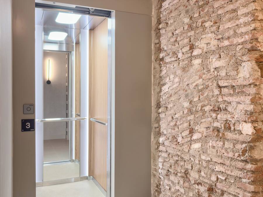 A hallway with a modern elevator, a brick wall, and a door leading to a room.