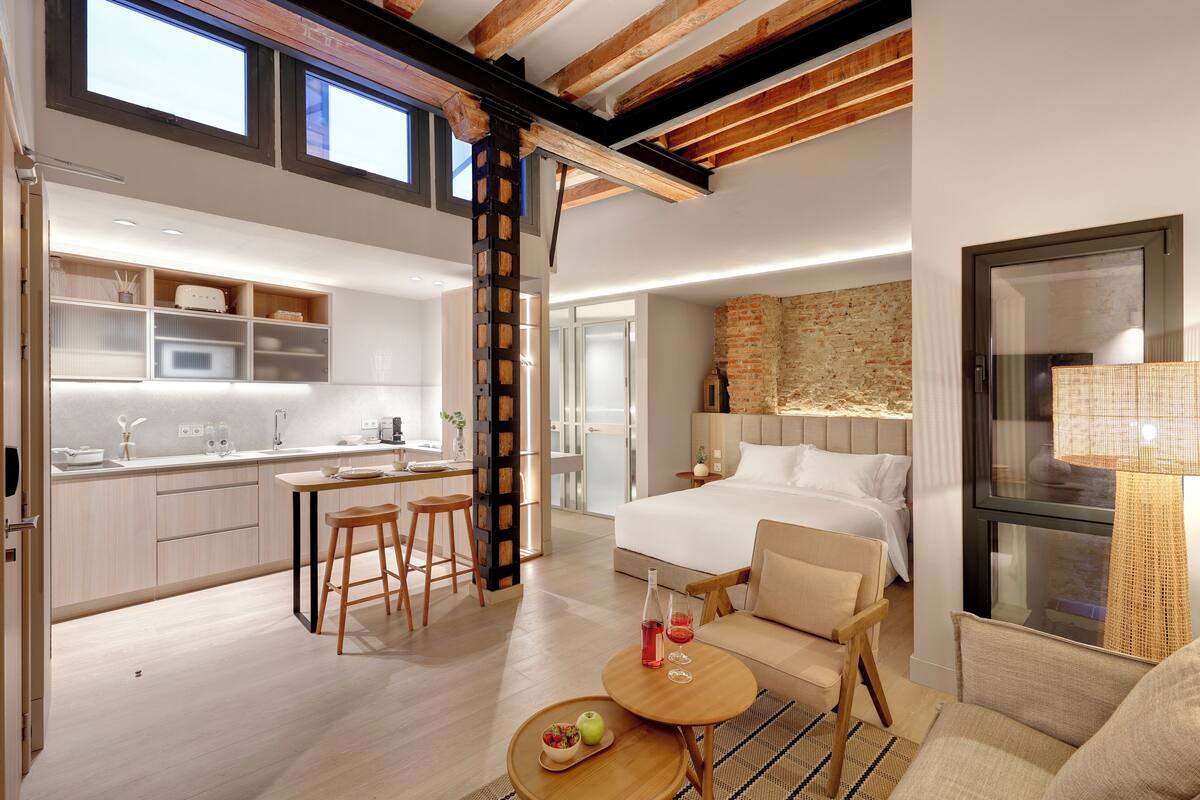 A modern hotel room with a large bed, a dining area with a kitchen, and a view of the city.