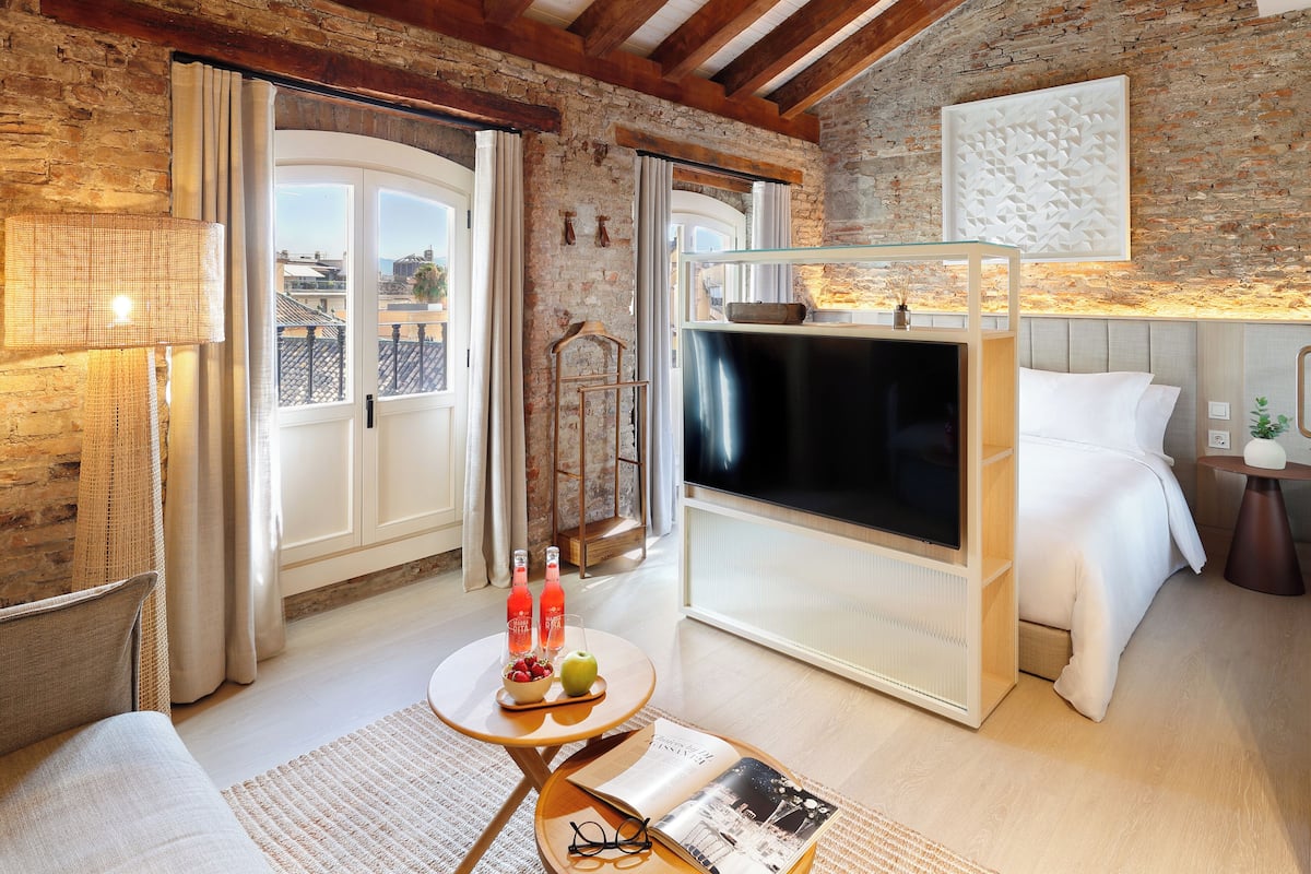 A cozy room with a TV, a bed, a table with fruit and drinks, and a view of the city through large windows.
