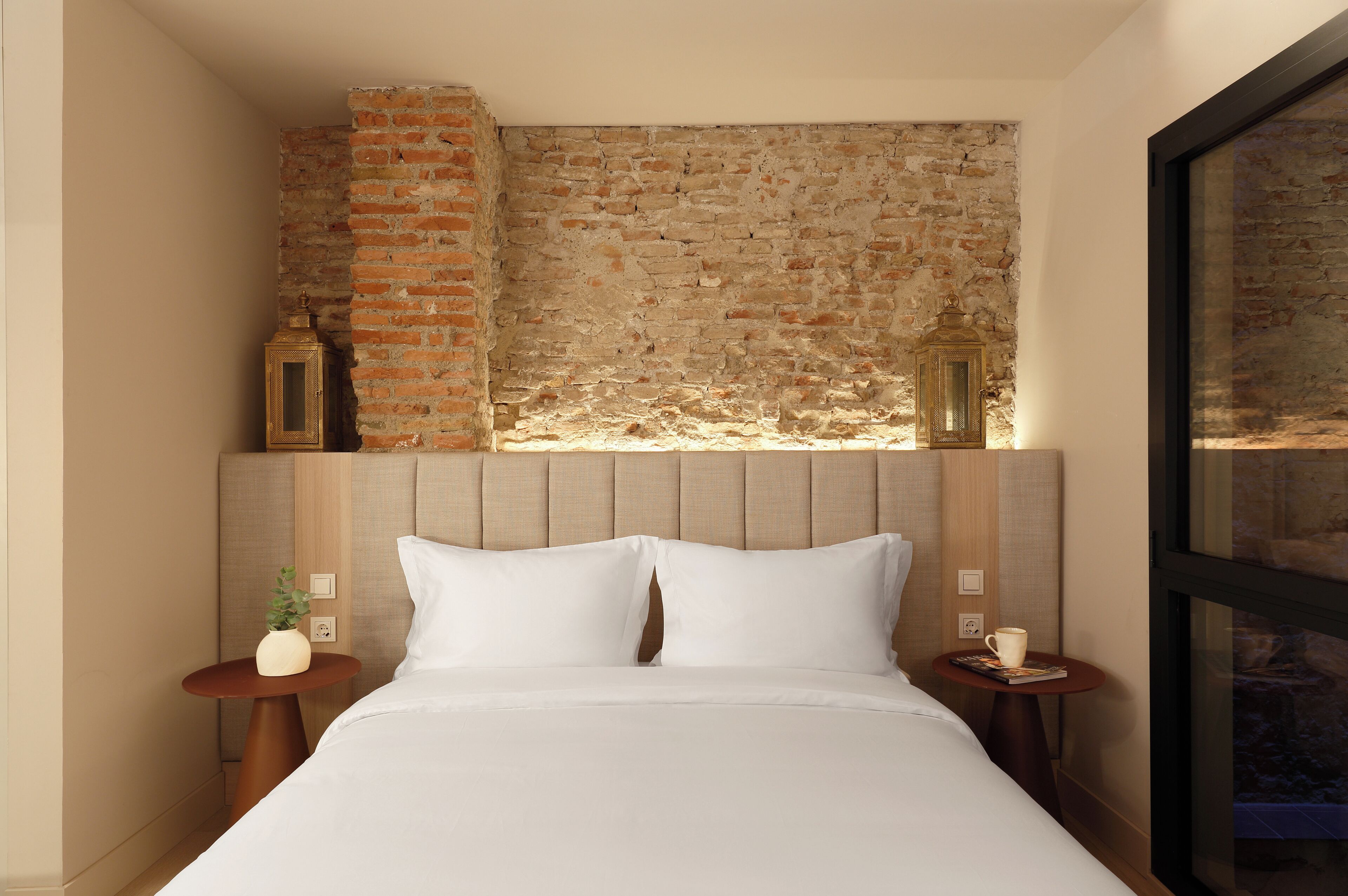 Een hotelkamer met een bakstenen muur, een bed met witte lakens, twee nachtkastjes en twee wandlampen.
