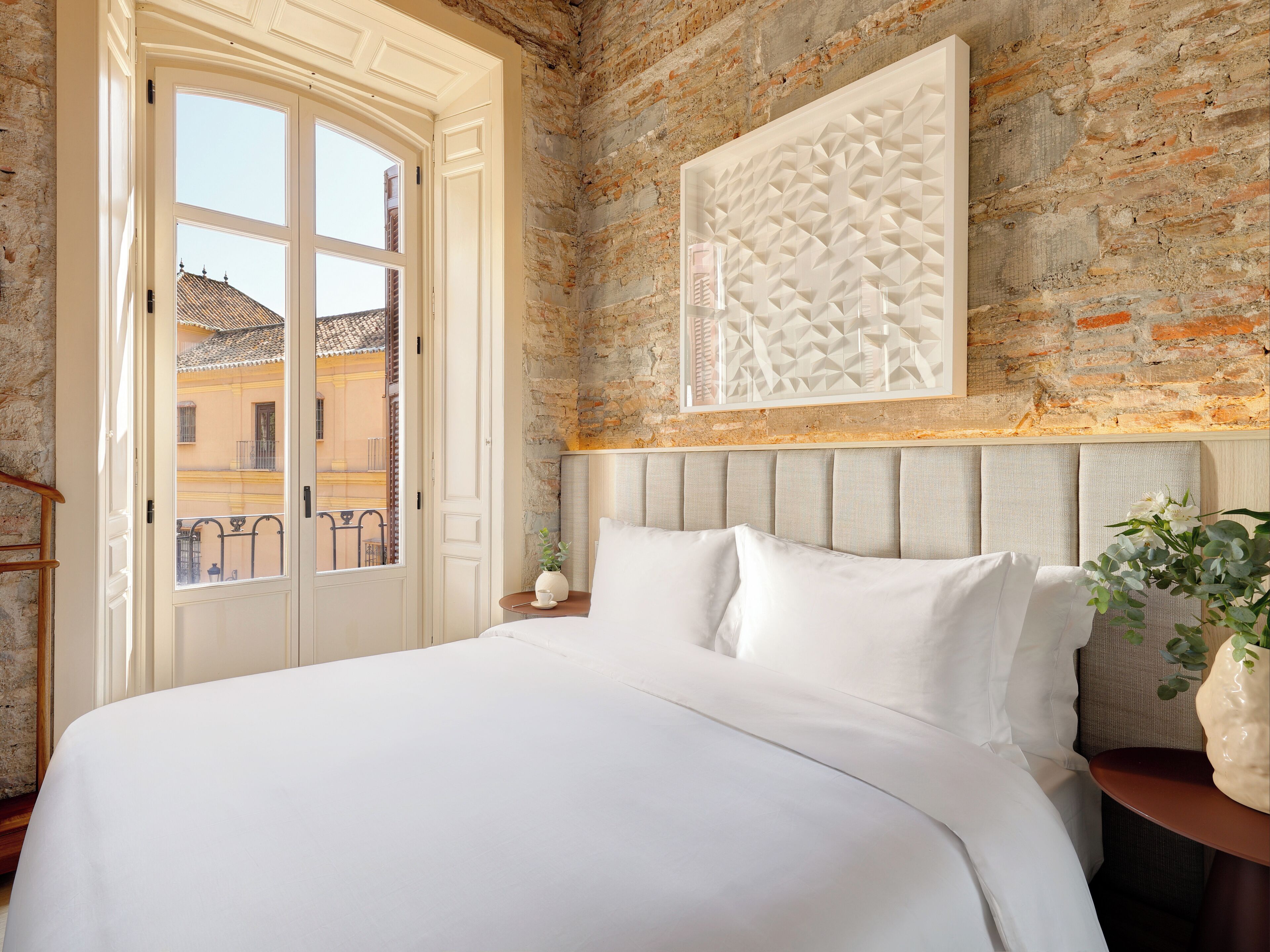 A bedroom with a large bed, a brick wall, a window with a view of buildings, and a decorative wall piece.