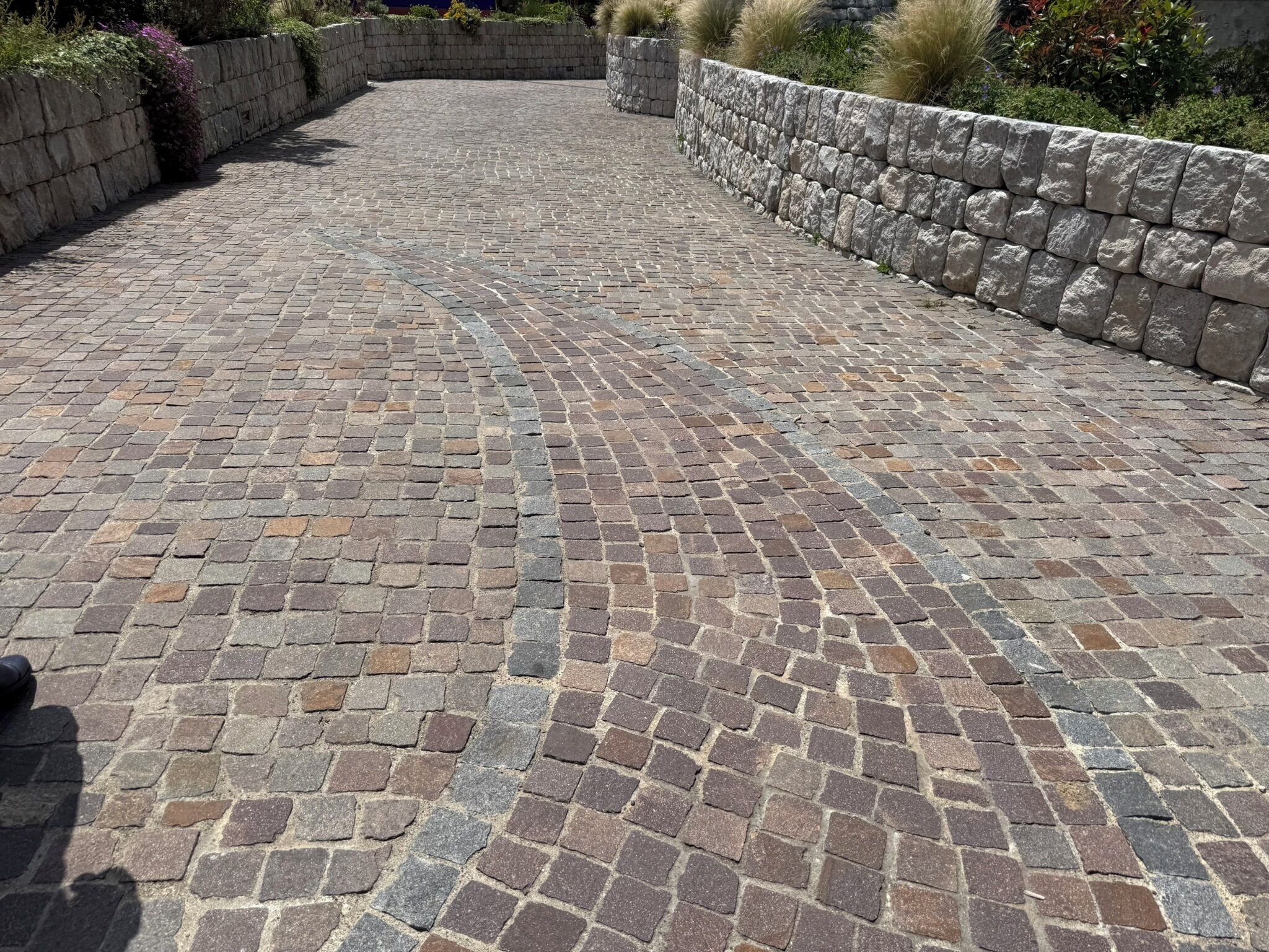 A paved pathway with stone walls on either side, leading to a landscaped area.