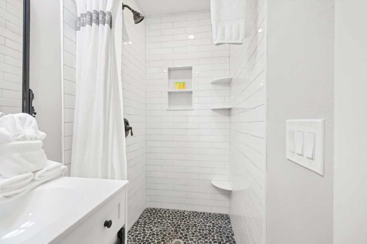 Une salle de bain avec des murs carrelés de blanc, une douche avec un rideau, un lavabo avec une pile de serviettes et une étagère contenant des articles de toilette.