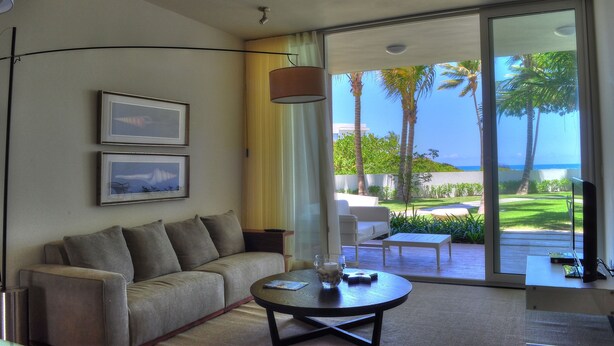 A modern living room with a sofa, a coffee table, and a TV. There are two framed pictures on the wall. The room has a large sliding glass door leading to a balcony with palm trees and a view of the ocean. - Oceanfront 1BR en Ultravioleta Cabarete. Wifi Rápido, Balcón & Parking (Cabarete)