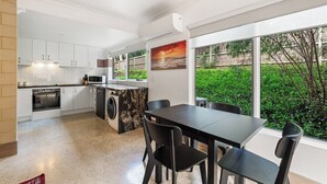 Eine moderne Küche mit weißen Schränken, einem eingebauten Backofen und einer schwarzen Arbeitsplatte. Es gibt eine Essecke mit Tisch und Stühlen, und ein großes Fenster mit Blick auf Grünflächen.