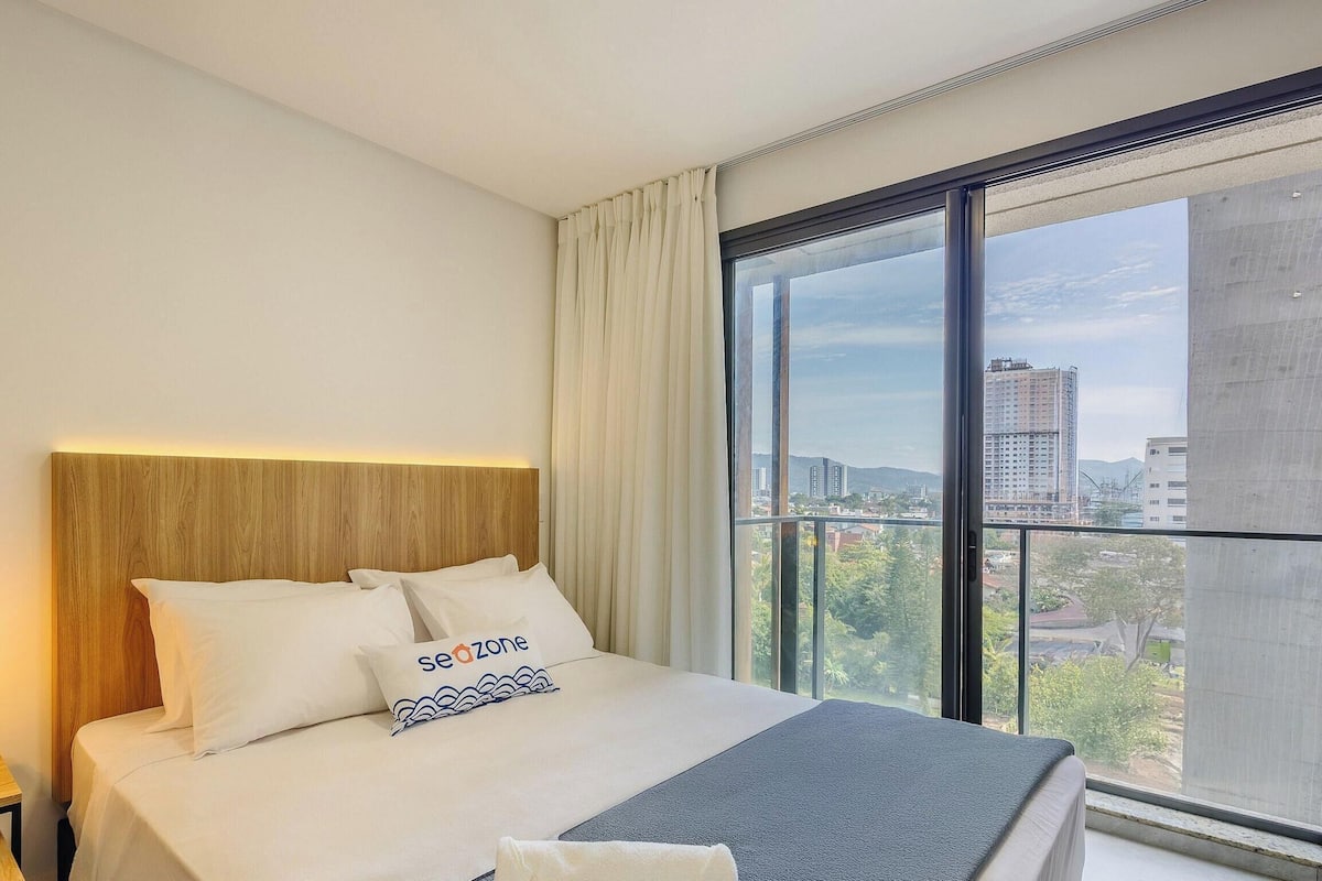Quarto de hotel com uma grande janela, uma cama com roupa de cama branca, uma cabeceira de madeira e uma mesa de cabeceira com abajur.