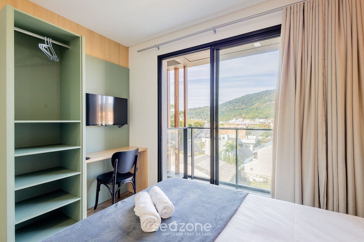 Quarto de hotel com cama, escrivaninha, cadeira, televisão e uma janela grande com cortinas.