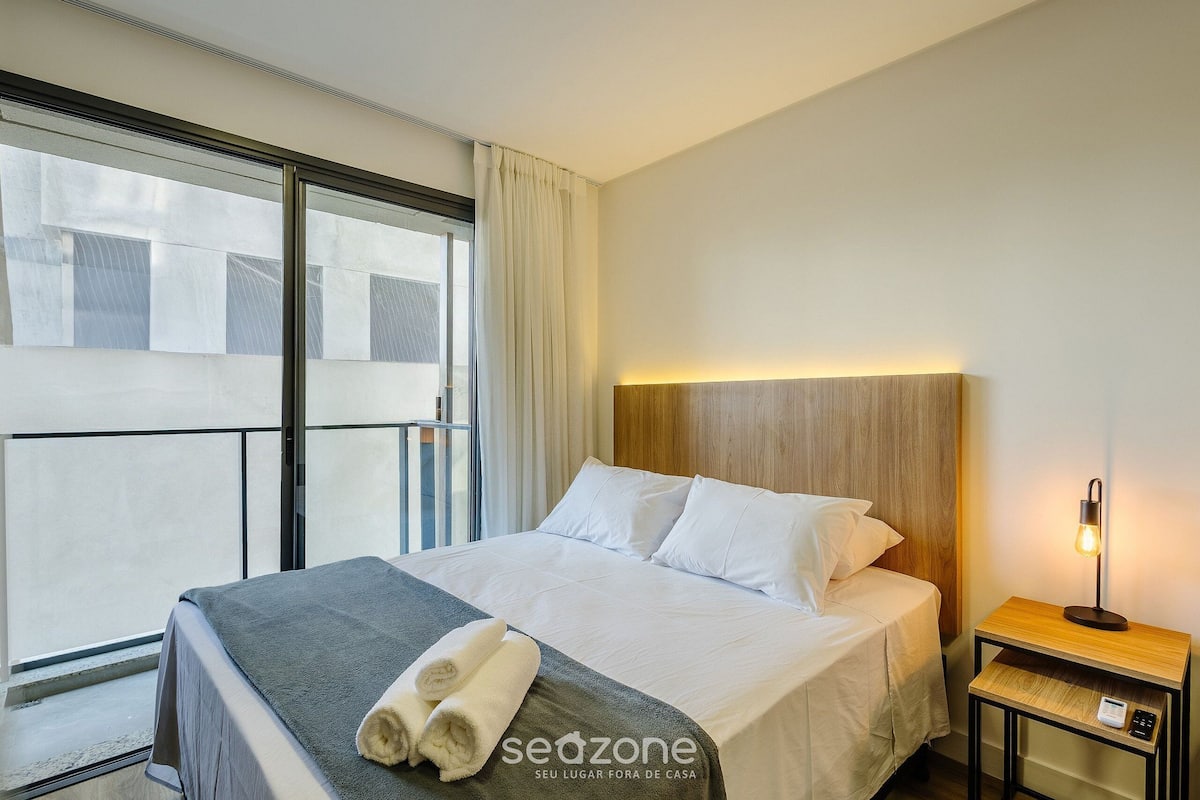 Quarto de hotel com uma cama, mesa de cabeceira com abajur, janela com cortinas e vista para edifícios.