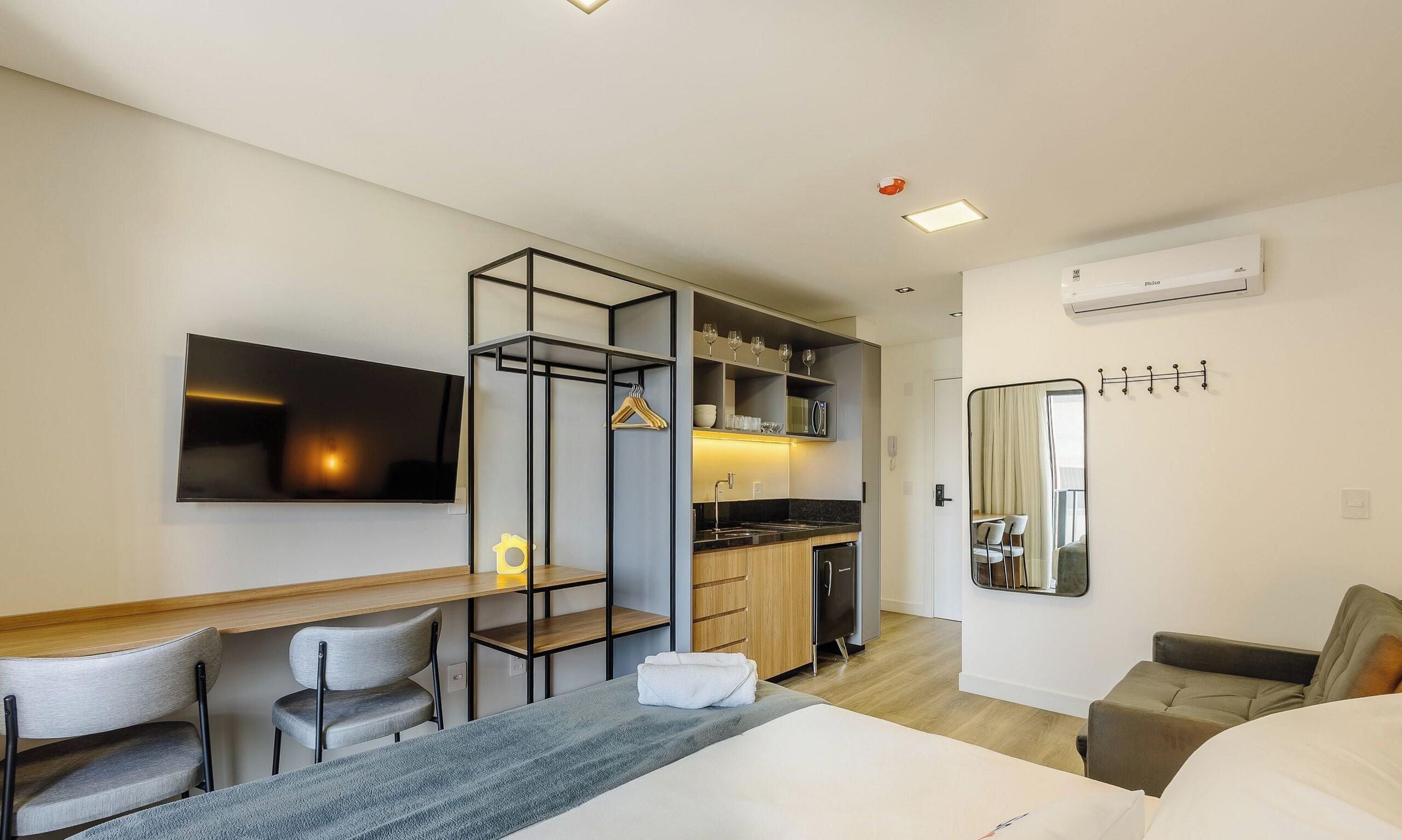 A modern hotel room with a bed, a desk, a chair, a kitchenette, and a bathroom.
