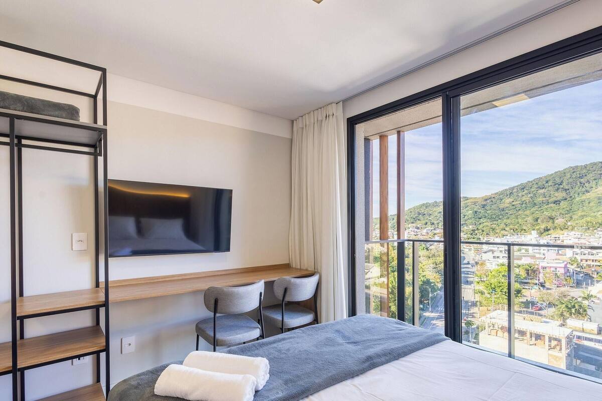 Quarto de hotel com cama, televisão na parede, secretária com cadeiras e uma grande janela com vista para a cidade.