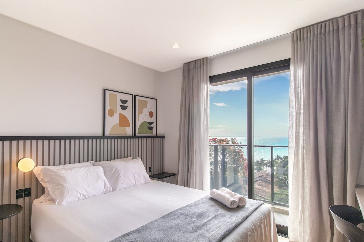 Quarto de hotel com cama, mesa de cabeceira, mesa e uma janela grande com cortinas.