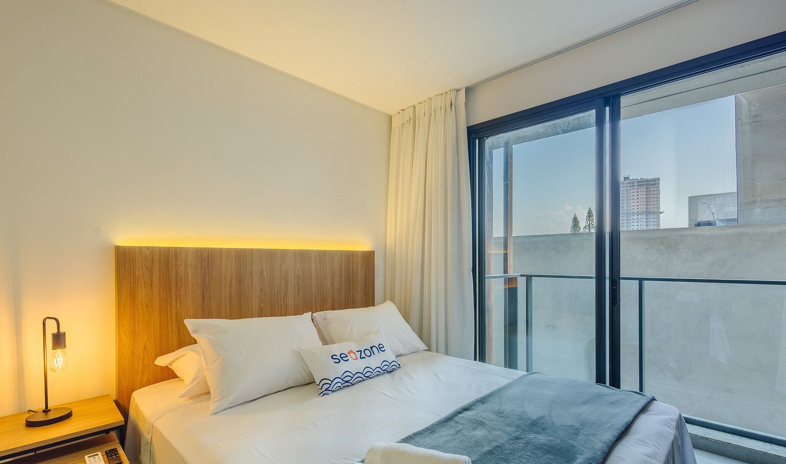 A hotel room with a bed, a nightstand, a lamp, a window with a view of buildings, and a curtain.