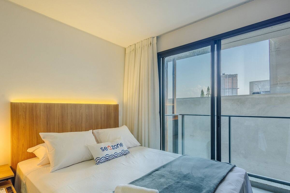 Quarto de hotel com cama, mesa de cabeceira, luminária, janela com vista para prédios e cortina.