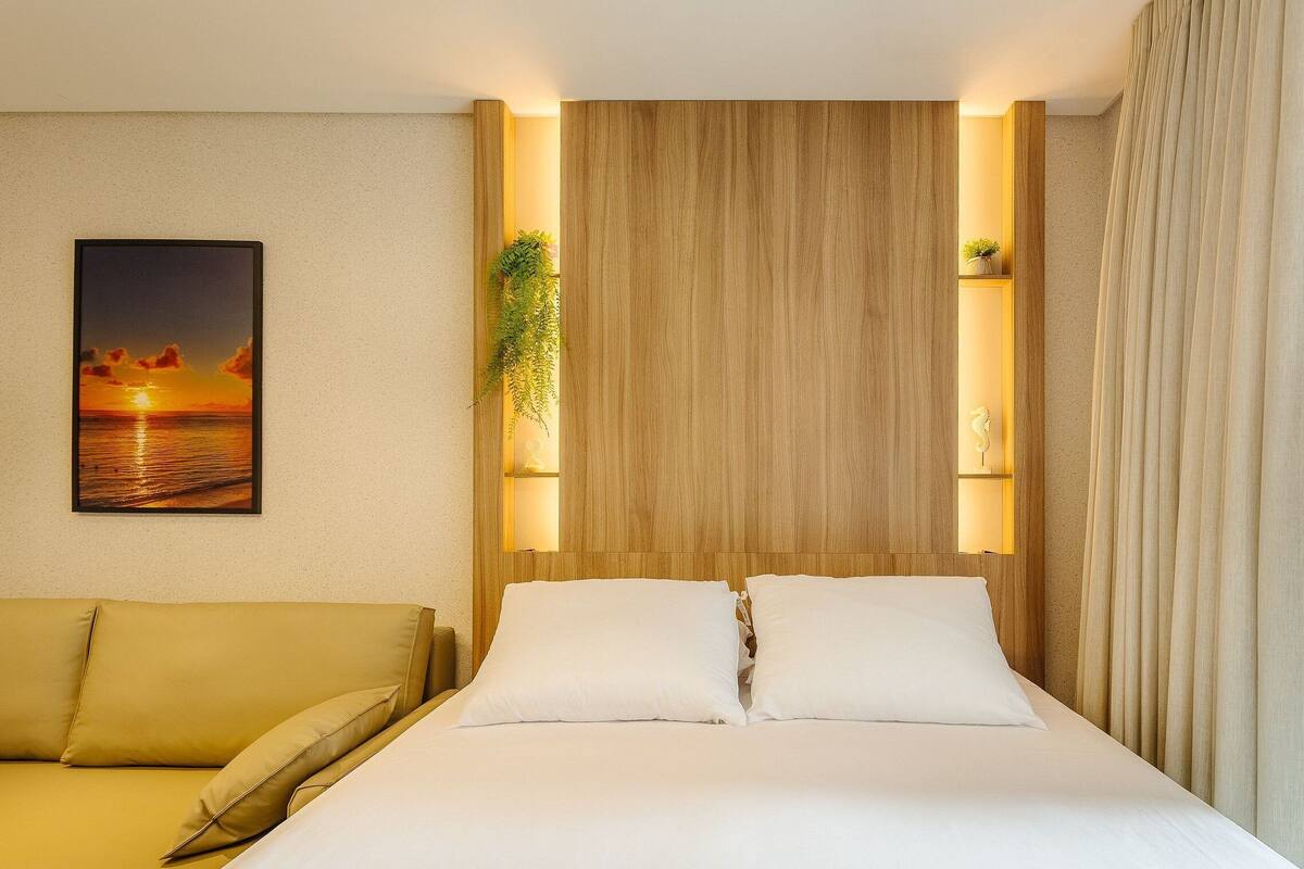 Quarto de hotel com uma cama, um sofá, prateleira na parede com uma planta e um quadro com uma imagem do pôr do sol.