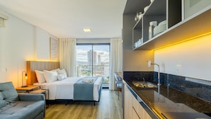 A modern hotel room with a bed, a sofa, a desk, a large window, and a kitchen area with a sink and cabinets. - Studios in a Seaside Building, Close to Beto Carreiro at Penha Spot - Psob (Penha)
