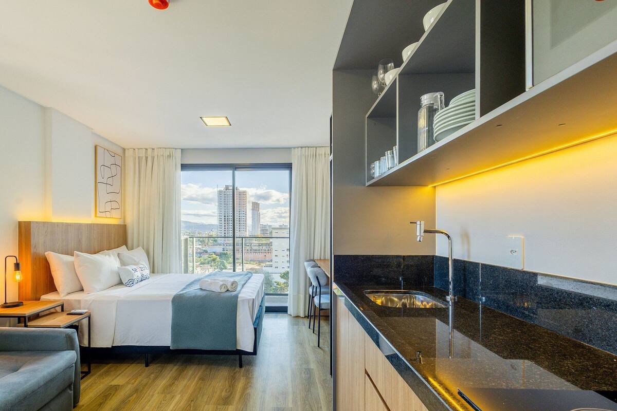 Quarto de hotel moderno com cama, sofá, escrivaninha, janela grande e área de cozinha com pia e armários.