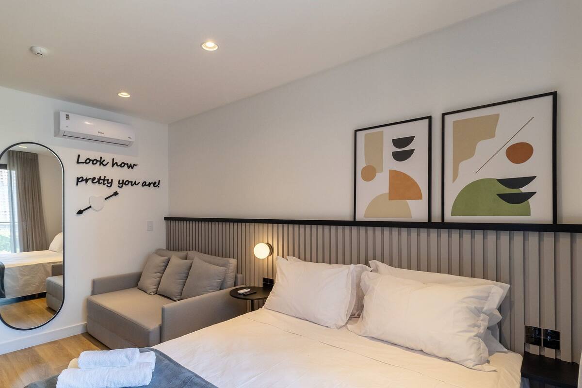 Quarto de hotel moderno com cama, área de estar, espelho e obras de arte nas paredes.