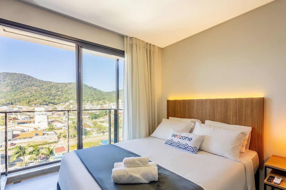 Quarto de hotel com uma janela grande, cama com roupa de cama branca, cabeceira de madeira e mesa de cabeceira com abajur.