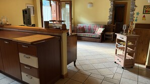 A kitchen with wooden cabinets, a countertop, and a tiled floor. There is a sofa and a dining table in the background.
