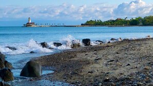 A rocky beach with waves crashing onto the shore, a lighthouse in the distance, and a small island with buildings and palm trees.